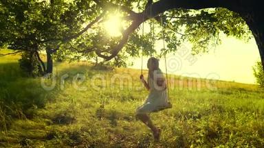 快乐的女孩留着长发，在树枝上荡秋千，在金色夕阳的明亮光线下微笑。 慢动作。